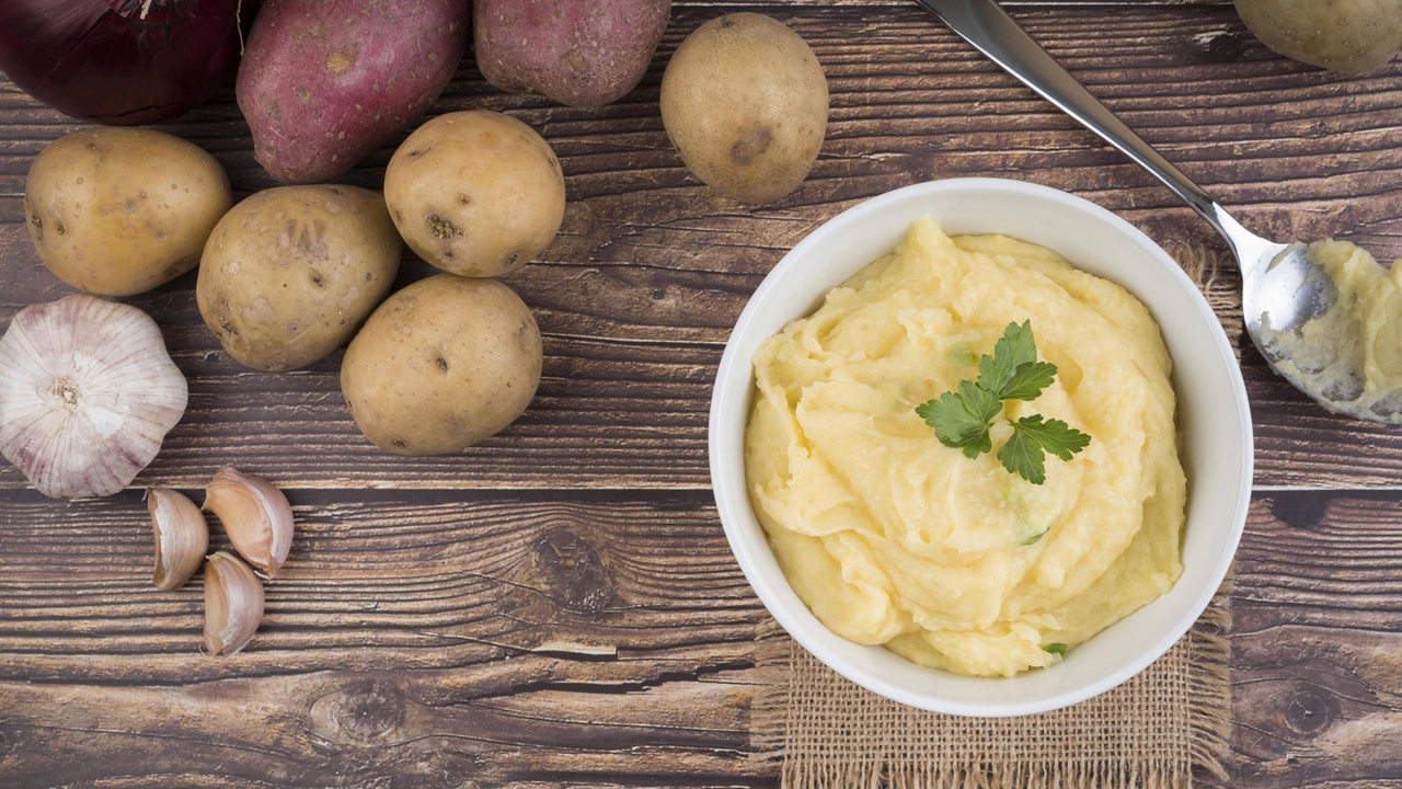 Eine Schüssel mit Kartoffelpüree. Daneben liegen ungekochte Kartoffeln und eine Knoblauchzehe.